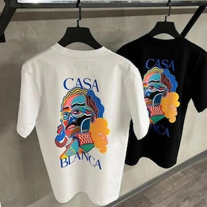 May include: Two t-shirts, one white and one black, hanging on a rack. Each shirt features a colorful graphic of a face with the words "CASA BLANCA" in blue and yellow. The design is printed on the back of the shirts.