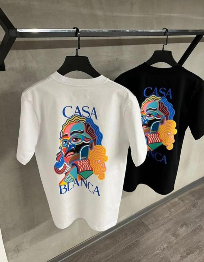 May include: Two t-shirts, one white and one black, hanging on a rack. Each shirt features a colorful graphic of a face with the words "CASA BLANCA" in blue and yellow. The design is printed on the back of the shirts.