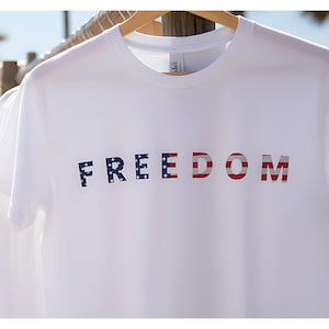 May include: A white t-shirt with the word "FREEDOM" printed across the front. The letters are in a patriotic design, with the letters "F", "R", "E", "E", and "D" featuring a blue background with white stars, and the letters "O" and "M" featuring red and white stripes.