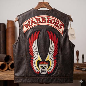 Può includere: Un gilet in pelle marrone con una grande toppa con la scritta "WARRIORS" in rosso e bianco. Sotto, un teschio con ali rosse, bianche e gialle è ricamato sul retro. Il gilet è appeso a una gruccia di legno.