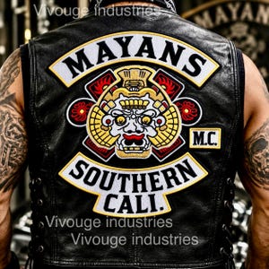 May include: A black leather vest featuring a large embroidered patch on the back. The patch displays the text MAYANS, SOUTHERN CALI., and M.C. with a central graphic of a stylized face in yellow, red, and white. The vest has side lacing details.