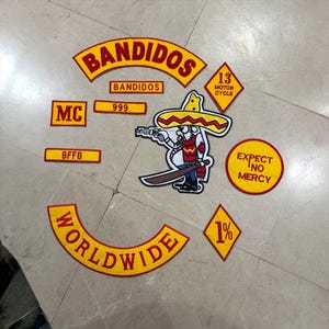 Puede incluir: Un conjunto de parches bordados en amarillo y rojo. Incluye un rocker grande de Bandidos, un personaje de dibujos animados con sombrero que sostiene una pistola y una espada, un rombo de 13 Motor Cycle, una tira 999, MC, BFFB, un rocker Worldwide, un rombo de 1% y un parche circular con el texto EXPECT NO MERCY.