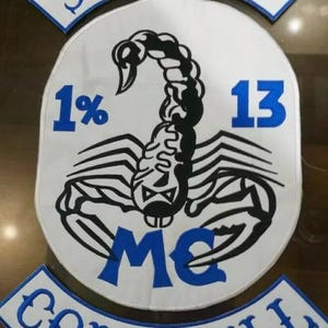 May include: A white patch with a black scorpion design, the text "SCORPIO", "1%", "13", "MC", and "CORNWALL" in blue. The patch is likely for a motorcycle club.