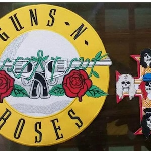 May include: Embroidered patches featuring the Guns N' Roses logo and a cross with skull designs. The round patch has a yellow border with the band's name and a gun and roses graphic. The cross patch has five skull designs.