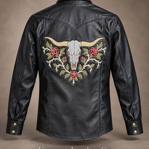 May include: A black leather jacket featuring a large embroidered design on the back. The embroidery depicts a white longhorn skull surrounded by red roses and green vines. The jacket has a collar, long sleeves with gold-tone buttons, and a yoke.