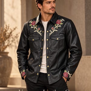 May include: A black leather shirt jacket featuring embroidered red rose motifs with green leaves on the chest and cuffs. The garment includes a pointed collar, two chest pockets with flaps, and gold-toned metal snap buttons down the front.