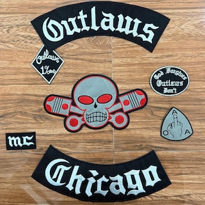 May include: A selection of black and white embroidered patches, including the words "Outlaws" and "Chicago". The patches feature a skull with red eyes and a middle finger graphic. These patches are for a motorcycle club.