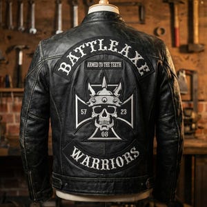 Op de afbeelding: Zwarte leren jas met een grote patch op de rug. De patch toont een schedel met een kroon, een kruis en de woorden "BATTLEAXE WARRIORS" en "ARMED TO THE TEETH". De jas wordt getoond op een mannequin in een werkplaats.