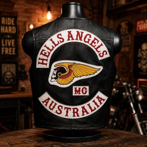 May include: A black leather vest featuring a large embroidered patch on the back. The patch displays the text HELLS ANGELS in red on a white curved background, a winged skull emblem in yellow and red, the letters MG, and the text AUSTRALIA in red.