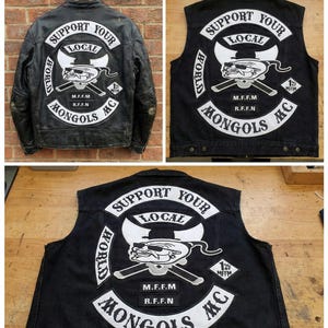 Puede incluir: Chaqueta de cuero negra y chalecos vaqueros con el logo "Mongols MC". El logo incluye un personaje de dibujos animados con gafas de sol, espadas cruzadas y el texto "Support Your Local". La chaqueta y los chalecos se muestran sobre una superficie de madera.
