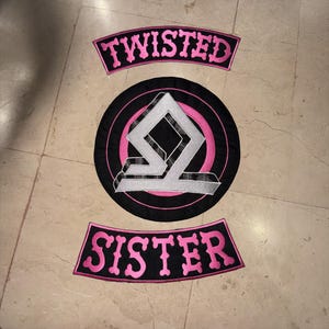 Pode incluir: Um conjunto de três emblemas bordados com um esquema de cores preto e rosa. Os emblemas curvos superior e inferior exibem o texto TWISTED e SISTER em letras cor-de-rosa. O emblema circular central apresenta um símbolo geométrico prateado.