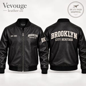 Op de afbeelding: Een zwart leren bomberjack met een ritssluiting aan de voorzijde en geribbelde boorden bij de kraag, mouwen en zoom. De voorzijde toont 'EST. 1992' en 'BROOKLYN HERITAGE CO.' in witte tekst, met 'BK' op de mouw. Achterop staat 'BROOKLYN CITY HERITAGE'.