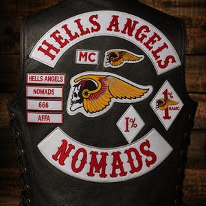 Könnte beinhalten: Eine schwarze Lederweste mit gestickten Aufnähern, darunter der Schriftzug HELLS ANGELS in einem weißen Bogen, ein geflügeltes Totenkopf-Emblem, der Text NOMADS und rautenförmige 1%-Aufnäher. Die Weste hat eine seitliche Schnürung und weitere rechteckige Textaufnäher.