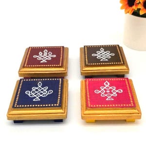 May include: A set of four square coasters with gold-painted edges and bases. Each coaster features a distinct solid color surface—maroon, dark brown, navy blue, and magenta—adorned with a unique white geometric line pattern and dotted border.