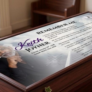 May include: A polished wooden casket with a printed memorial. The memorial features a portrait of a man, the name "Keith Joyner", and the words "Remember Me" with a poem. White roses and greenery decorate the top of the casket.