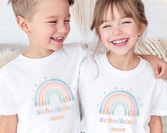 May include: Two children wearing white t-shirts with a pastel rainbow design and the text "Schulkind 2026". The shirts are paired with blue jeans. The children are smiling.
