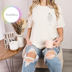 May include: Cream-colored t-shirt with a floral design and the word "Mama" printed in black. The shirt also features the text "She believed she could so she did." The model is holding a pink mug.