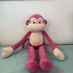 Peut inclure: Une peluche singe avec un corps rose, de longs bras et jambes, et un visage et des mains crème. Le singe a une expression souriante et est posé sur une surface bleu clair.