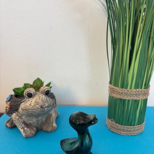 Peut inclure: Une petite figurine de canard en céramique bleu-vert foncé avec une finition brillante. Le canard est posé sur une surface bleu vif, avec un pot de grenouille et un vase d'herbe décorative en arrière-plan. Le pot de grenouille est gris et contient une plante succulente.