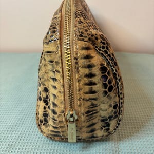 Peut inclure: Petite trousse de maquillage ovale avec un motif peau de serpent dans les tons beige, marron et noir. Le sac est dot&eacute; d'une fermeture &eacute;clair dor&eacute;e qui longe le dessus et d'un petit curseur de fermeture &eacute;clair en m&eacute;tal. Le sac est fait d'une mati&egrave;re textur&eacute;e.