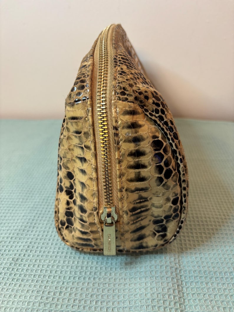 Peut inclure: Petite trousse de maquillage ovale avec un motif peau de serpent dans les tons beige, marron et noir. Le sac est dot&eacute; d'une fermeture &eacute;clair dor&eacute;e qui longe le dessus et d'un petit curseur de fermeture &eacute;clair en m&eacute;tal. Le sac est fait d'une mati&egrave;re textur&eacute;e.