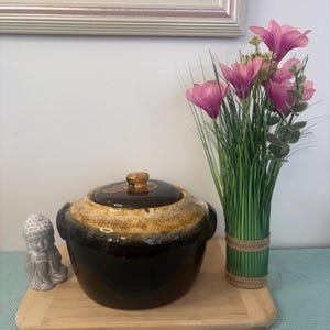 Peut inclure: Un plat à casserole en céramique marron foncé avec un couvercle, avec un bord doré et beige. Le plat est posé sur un plateau en bois, à côté d'une petite statue de Bouddha et d'un vase de fleurs roses et d'herbe verte.