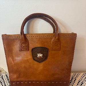 Puede incluir: Un bolso tote de cuero marrón con detalles cosidos y un emblema decorativo. El bolso tiene dos asas y una forma rectangular. El emblema tiene forma de escudo con un animal de color dorado.
