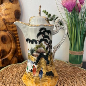 Puede incluir: Una cafetera de porcelana con un paisaje pintado a mano que incluye figuras con vestimenta tradicional, pinos negros y una base amarilla. La pieza presenta un borde festoneado, un asa curva con detalles dorados y una tapa a juego con remate.