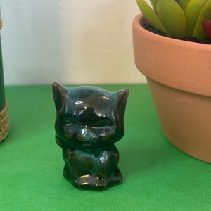 May include: A small, ceramic cat figurine in shades of dark green and brown. The cat is sitting with its eyes closed, and the glaze gives it a glossy finish. The figurine is on a green surface, with a terracotta pot and a plant in the background.