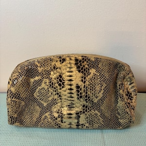 Peut inclure: Une trousse de maquillage beige et noire à motif peau de serpent avec une fermeture éclair dorée. Le sac a une forme arrondie et une surface texturée, avec un panneau central d'écailles beige clair.