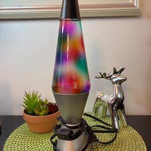 Op de afbeelding: Een lavalamp met een conische glazen houder met veelkleurige vloeistof in tinten rood, groen en paars. De lamp heeft een zwarte dop en een zilverkleurige metalen voet met een bijbehorend zwart netsnoer en een ingebouwde schakelaar.