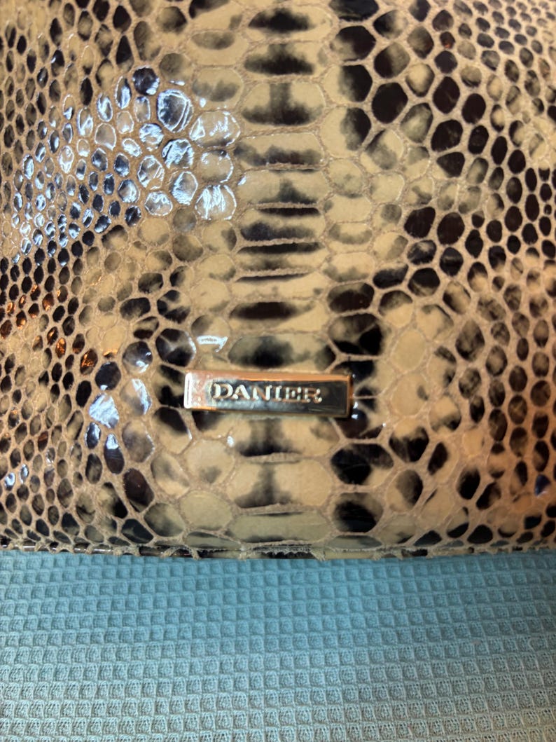 Peut inclure: Gros plan sur un sac &agrave; main avec un motif peau de serpent dans les tons beige, noir et marron. Le sac &agrave; main a une finition brillante et une plaque signal&eacute;tique en m&eacute;tal argent&eacute; qui indique "DANIER".