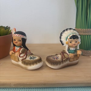 May include: A pair of vintage ceramic figurines depicting Native American children. Each figurine is seated in a brown shoe with floral accents. One wears a headdress, the other a headband. The figurines are on a wooden surface.