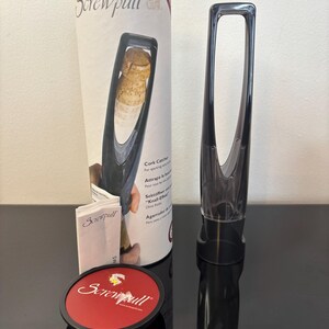 May include: A black Screwpull cork catcher and a clear wine opener are displayed next to a red and black coaster. The packaging includes the text "Screwpull" and "Cork Catcher". The wine opener is designed for sparkling wine.