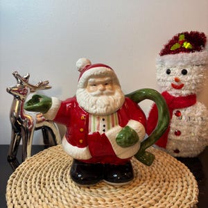 May include: A ceramic teapot shaped like Santa Claus, featuring a red coat with white trim, a green handle, and green mittens. Santa has a white beard, a red hat with a white pom-pom, and black boots. The teapot is decorated with a striped vest.