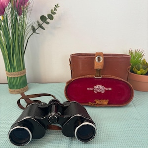 Pode incluir: Binóculos pretos com uma correia de couro castanho e detalhes prateados, ao lado de um estojo de couro castanho com interior de veludo vermelho. O estojo está aberto, mostrando o texto "BAKER STANDARD TESTED". Flores cor-de-rosa e vegetação no fundo.