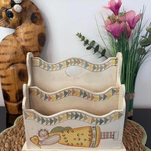 May include: A cream-coloured wooden letter holder featuring three tiered compartments with wavy edges. Each tier is decorated with a painted border of small triangles in muted tones. The front panel displays a painted figure in a yellow dress.