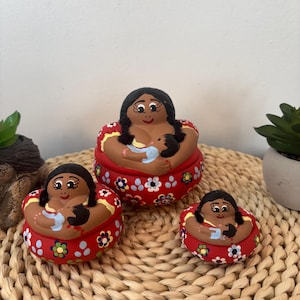 Puede incluir: Tres recipientes redondos de cerámica roja con tapa de diferentes tamaños, cada uno con una figura pintada a mano de cabello negro que sostiene a un bebé envuelto en una manta azul claro. La base de cada recipiente tiene un diseño floral.