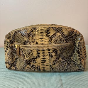 Peut inclure: Une trousse de maquillage rectangulaire &agrave; motif peau de serpent, avec une fermeture &eacute;clair et une tirette dor&eacute;es. Le sac pr&eacute;sente un m&eacute;lange de couleurs beige, noir et cr&egrave;me, probablement en cuir. Il a un dessus arrondi et une base plate.