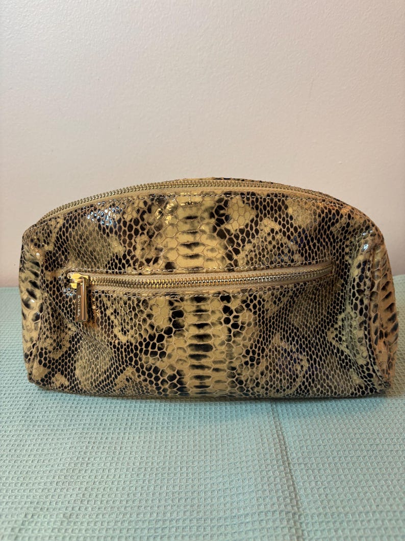 Peut inclure: Une trousse de maquillage rectangulaire &agrave; motif peau de serpent, avec une fermeture &eacute;clair et une tirette dor&eacute;es. Le sac pr&eacute;sente un m&eacute;lange de couleurs beige, noir et cr&egrave;me, probablement en cuir. Il a un dessus arrondi et une base plate.