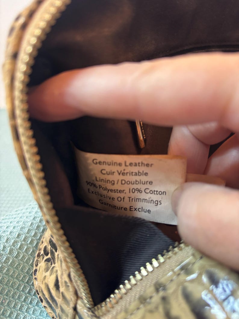 Peut inclure: Gros plan sur un petit sac &agrave; main &agrave; motifs avec une fermeture &eacute;clair. L'int&eacute;rieur du sac est visible, ainsi qu'une &eacute;tiquette indiquant "Genuine Leather, Cuir V&eacute;ritable, Lining/Doublure, 90% Polyester, 10% Cotton, Exclusive Of Trimmings, Garniture Exclue."