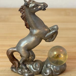 May include: A silver-colored figurine of a rearing horse with a golden orb. The horse is detailed with a brown mane and tail, standing on a cloud-like base. The orb is translucent and sits beside the horse.