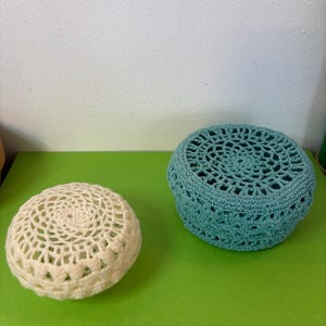 Peut inclure: Deux objets faits à la main au crochet. L'un est une petite pièce ronde, blanc cassé, avec un motif ajouré. L'autre est un objet rond, plus grand, de couleur sarcelle, avec un motif ajouré similaire. Les deux objets sont sur une surface verte.