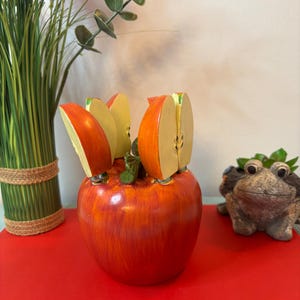 May include: A red apple-shaped knife holder with four apple slice-shaped knives. The knife holder is ceramic and has a glossy finish. The knives are made of wood and have a realistic appearance. Decorative kitchenware.