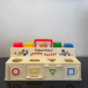 Peut inclure: Un jouet trieur de formes Fisher-Price vintage. Le jouet est de couleur crème avec une poignée rouge et comporte quatre blocs colorés : rouge, jaune, vert et bleu. Le trieur de formes comporte quatre ouvertures de formes : cercle, rectangle, triangle et carré.