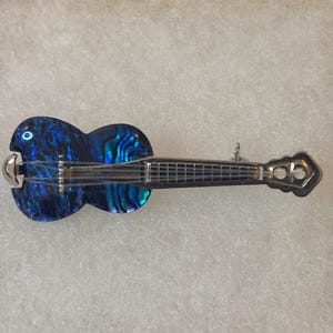 Peut inclure: Une broche en forme de guitare avec un corps incrusté de nacre bleue et noire irisée. Le manche, le chevalet et la tête sont fabriqués en métal argenté poli, avec de fines cordes métalliques courant le long de la touche.