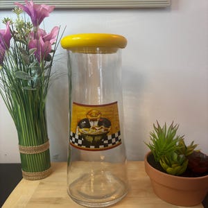 Peut inclure: Un bocal en verre transparent avec un couvercle jaune. Le bocal porte une étiquette décorative représentant un chef tenant une assiette de nourriture. L'étiquette est bordée d'un motif à carreaux noir et blanc. Le bocal est sur une planche à découper en bois.