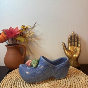 May include: A blue ceramic clog-shaped planter with a speckled finish, featuring a sculpted pink rose and a green leaf on the toe. The object has a hollow interior and a smooth, glossy glaze, presenting a classic footwear silhouette in a decorative form.