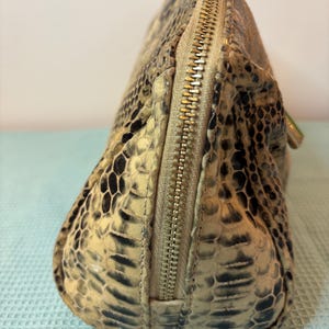 Peut inclure: Une trousse de maquillage beige et noire &agrave; motif peau de serpent avec une fermeture &eacute;clair dor&eacute;e. Le sac a une forme arrondie et une surface textur&eacute;e, avec une fermeture &eacute;clair sur le dessus. Le sac est fait d'un mat&eacute;riau qui ressemble &agrave; de la peau de serpent.