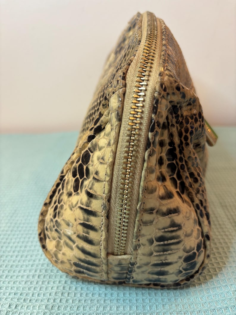 Peut inclure: Une trousse de maquillage beige et noire &agrave; motif peau de serpent avec une fermeture &eacute;clair dor&eacute;e. Le sac a une forme arrondie et une surface textur&eacute;e, avec une fermeture &eacute;clair sur le dessus. Le sac est fait d'un mat&eacute;riau qui ressemble &agrave; de la peau de serpent.